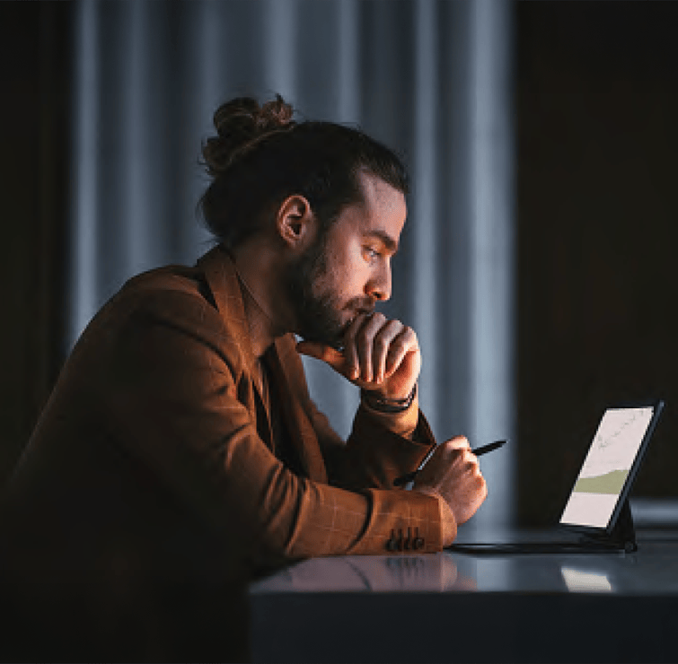 A man sits at a desk, resting his chin on his hand as he looks thoughtfully at a laptop