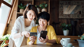 Joyful Asian senior mother and daughter shopping online with smartphone together and making payment with credit card at home