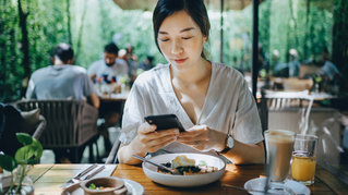young Asian woman using smartphone while having brunch in an outdoor restaurant surrounded by lush foliage and beautiful sunlight