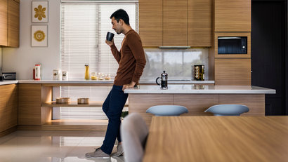 Young Asian man drinking coffee, leaning against modern kitchen island breakfast table with early morning sunlight 