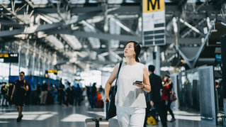 Ung asiatisk kvinnlig resenär med resväska kollar på smartphone medan hon går i den internationella flygplatshallen.
