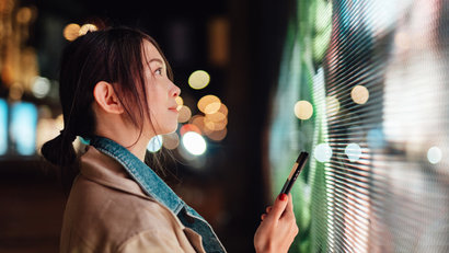 Young woman using mobile phone against led screen on city street at night connecting to the future