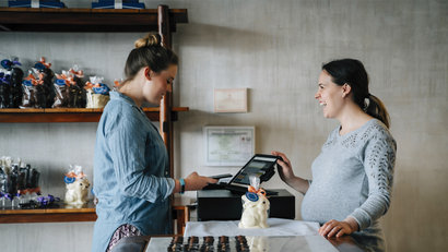 A smiling cashier accepts payment from a woman using her mobile phone in a small shop