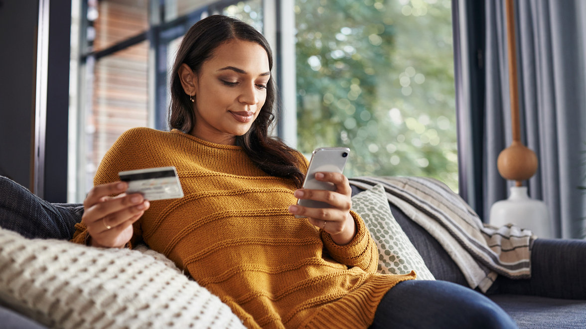 beautiful young woman using her cellphone and credit card while relaxing on a couch at home
