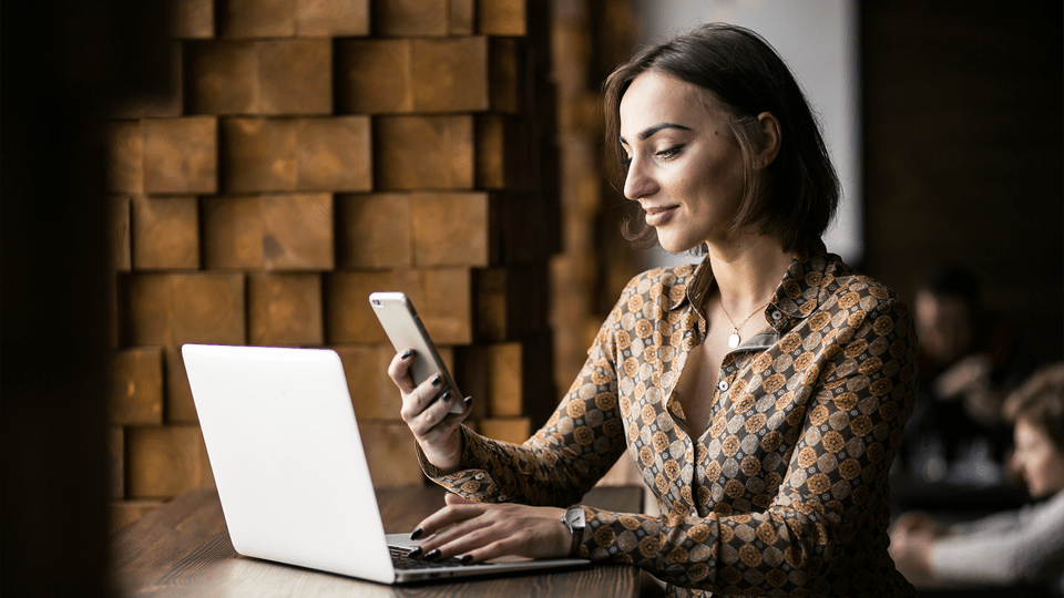 A woman typing on her laptop while looking at her phone smiling.