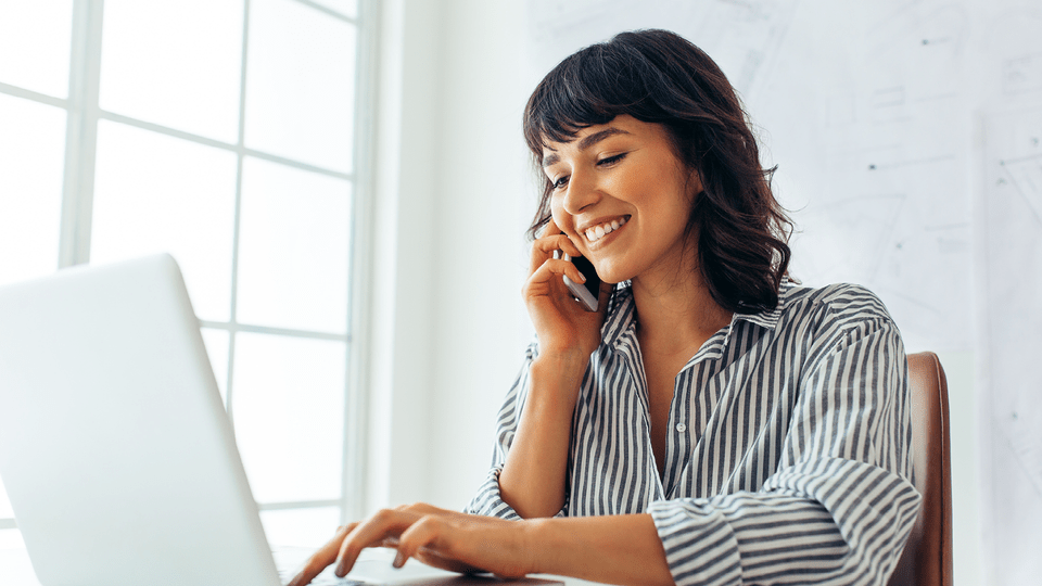 A woman going through her laptop while making a call smiling.