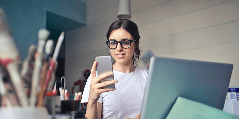  kvinne med briller ser på mobiltelefonen hennes foran en bærbar datamaskin