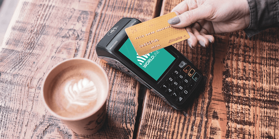 Hand betalar med kort över en worldline kortterminal med en kopp kaffe bredvid