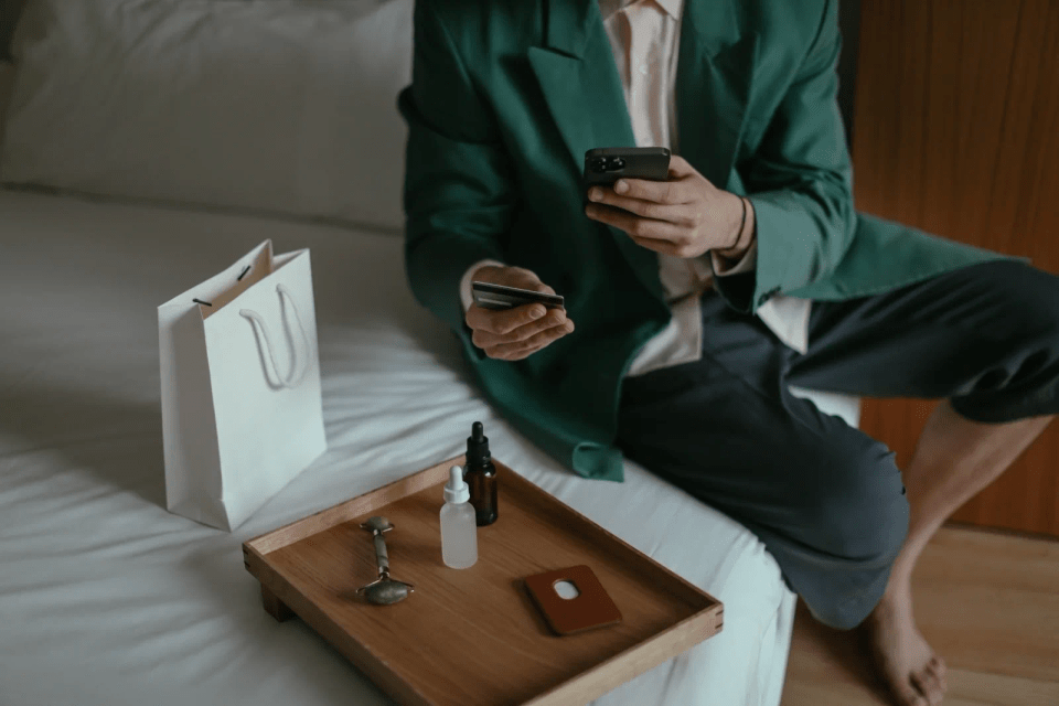 En mann sitter på sengekanten og betaler med kortet sitt via mobiltelefonen